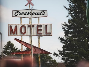 A vintage sign for the Crossroad's Motel, featuring large red lettering on a weathered metal background. The sign is mounted on rusty metal poles, with an adjacent arrow pointing towards the motel. Green trees are visible in the background against a partly cloudy sky.