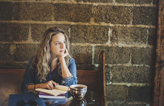A person sits at a table in a cozy, rustic setting, gazing thoughtfully into the distance. A notebook and a cup are placed in front, adding a sense of contemplation or study to the scene. The background features a textured brick wall, enhancing the warm, intimate atmosphere.