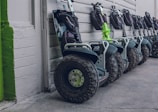 A row of Segway scooters is lined up against a wall. Each scooter is equipped with large, rugged tires and a handlebar with a small bag attached. The color scheme features mostly dark tones with some green accents.