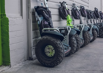 A row of Segway scooters is lined up against a wall. Each scooter is equipped with large, rugged tires and a handlebar with a small bag attached. The color scheme features mostly dark tones with some green accents.