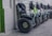 A row of Segway scooters is lined up against a wall. Each scooter is equipped with large, rugged tires and a handlebar with a small bag attached. The color scheme features mostly dark tones with some green accents.