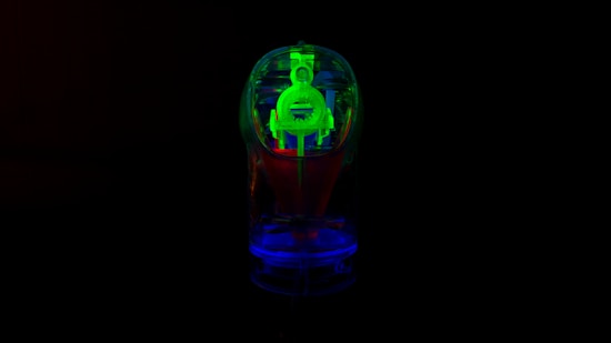 A neon-colored object, possibly a small transparent toy or gadget, with a glowing effect in green, blue, and red hues. The object is set against a completely black background, highlighting its luminous appearance.