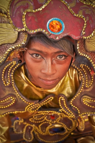 A person is adorned in an intricate, colorful costume with golden and red tones. The elaborate headpiece features a prominent orange gem and decorative patterns with small golden beads outlining it. The makeup is detailed and artistic, enhancing the visual impact of the outfit.