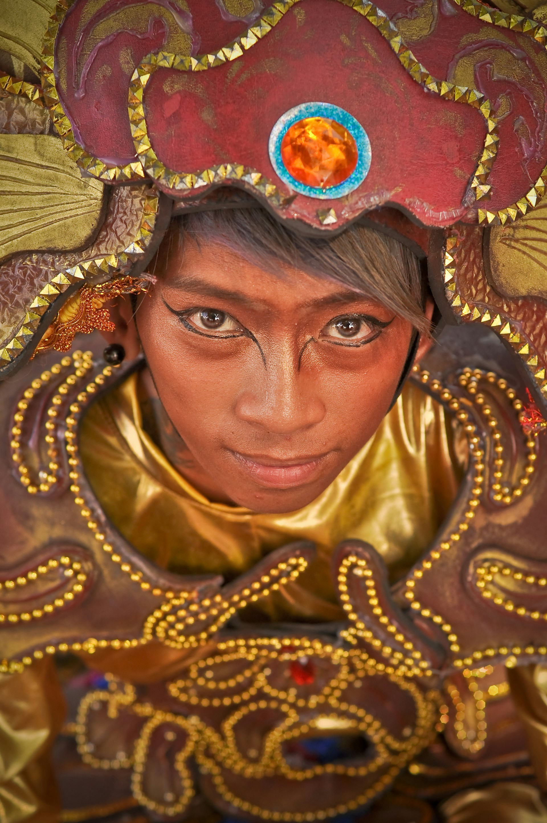 A person is adorned in an intricate, colorful costume with golden and red tones. The elaborate headpiece features a prominent orange gem and decorative patterns with small golden beads outlining it. The makeup is detailed and artistic, enhancing the visual impact of the outfit.