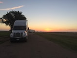 A large white semi-truck is parked on a rural road at dusk. The road is bordered by grass, and a substantial, leafy tree stands nearby, partially shading the truck. In the background, the sky transitions from light blue to warm orange hues as the sun sets on the horizon.