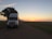 A large white semi-truck is parked on a rural road at dusk. The road is bordered by grass, and a substantial, leafy tree stands nearby, partially shading the truck. In the background, the sky transitions from light blue to warm orange hues as the sun sets on the horizon.