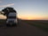 A large white semi-truck is parked on a rural road at dusk. The road is bordered by grass, and a substantial, leafy tree stands nearby, partially shading the truck. In the background, the sky transitions from light blue to warm orange hues as the sun sets on the horizon.
