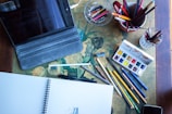 A workspace featuring art supplies, including colored pencils, a sketchbook, an iPad in a case, and a palette of watercolor paints arranged on a table with a floral pattern. The setup suggests a creative atmosphere with tools for drawing and painting.