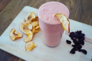 A creamy pink smoothie fills a glass, garnished with a dried apple slice. On a wooden board beside it, there are additional dried apple slices and a small pile of dark dried fruits, possibly berries.