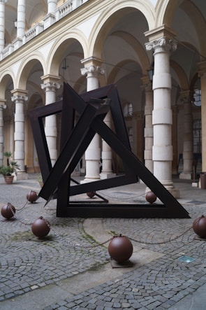 An abstract, geometric sculpture composed of dark metal beams forms overlapping triangular shapes. The structure is positioned outdoors in a courtyard with classical architecture, including stone columns and arches. Around the base of the sculpture, large metal balls connected by chains are placed on the cobblestone ground.