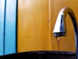A metal faucet is prominently positioned on the right, with a single droplet of water hanging from it. The background consists of vertical panels painted in vibrant blue and yellow hues.