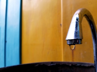 A metal faucet is prominently positioned on the right, with a single droplet of water hanging from it. The background consists of vertical panels painted in vibrant blue and yellow hues.
