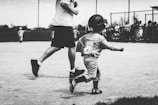 A child wearing a baseball uniform and helmet runs on a baseball field. The child has the number 6 on their jersey. An adult, partly visible and dressed casually, runs alongside the child. In the background, a group of people, possibly spectators, stands near a fence.