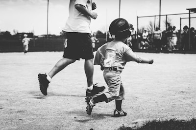 A child wearing a baseball uniform and helmet runs on a baseball field. The child has the number 6 on their jersey. An adult, partly visible and dressed casually, runs alongside the child. In the background, a group of people, possibly spectators, stands near a fence.