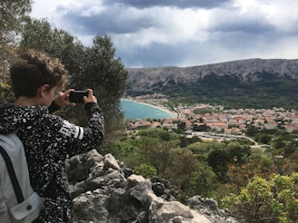 A person with a backpack is taking a photo with a smartphone from a rocky, elevated viewpoint. Below, a coastal town with red-roofed buildings and a curved beach is visible. The background consists of a mountain range under a cloudy sky.
