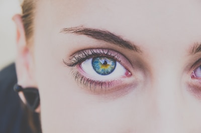 A close-up of a human face focusing on a bright, multicolored eye with striking hues of blue, green, and brown. The eyelashes are long and defined, while the eyebrow is well-groomed.