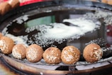 Golden-brown falafel balls with sesame seeds are lined up around the edge of a large metal frying pan filled with oil. The background shows a blurred mixture of colors, suggesting a vibrant, possibly outdoor setting.