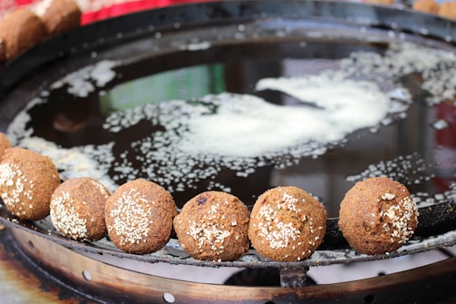Golden-brown falafel balls with sesame seeds are lined up around the edge of a large metal frying pan filled with oil. The background shows a blurred mixture of colors, suggesting a vibrant, possibly outdoor setting.