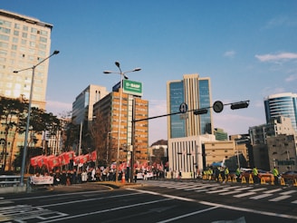 A group of people is gathered on a city street, some holding red flags and banners. The scene is set against a backdrop of tall, modern buildings under a clear blue sky. Traffic lights and a street sign are visible, and there are people wearing yellow vests, possibly security or police, lining the road.