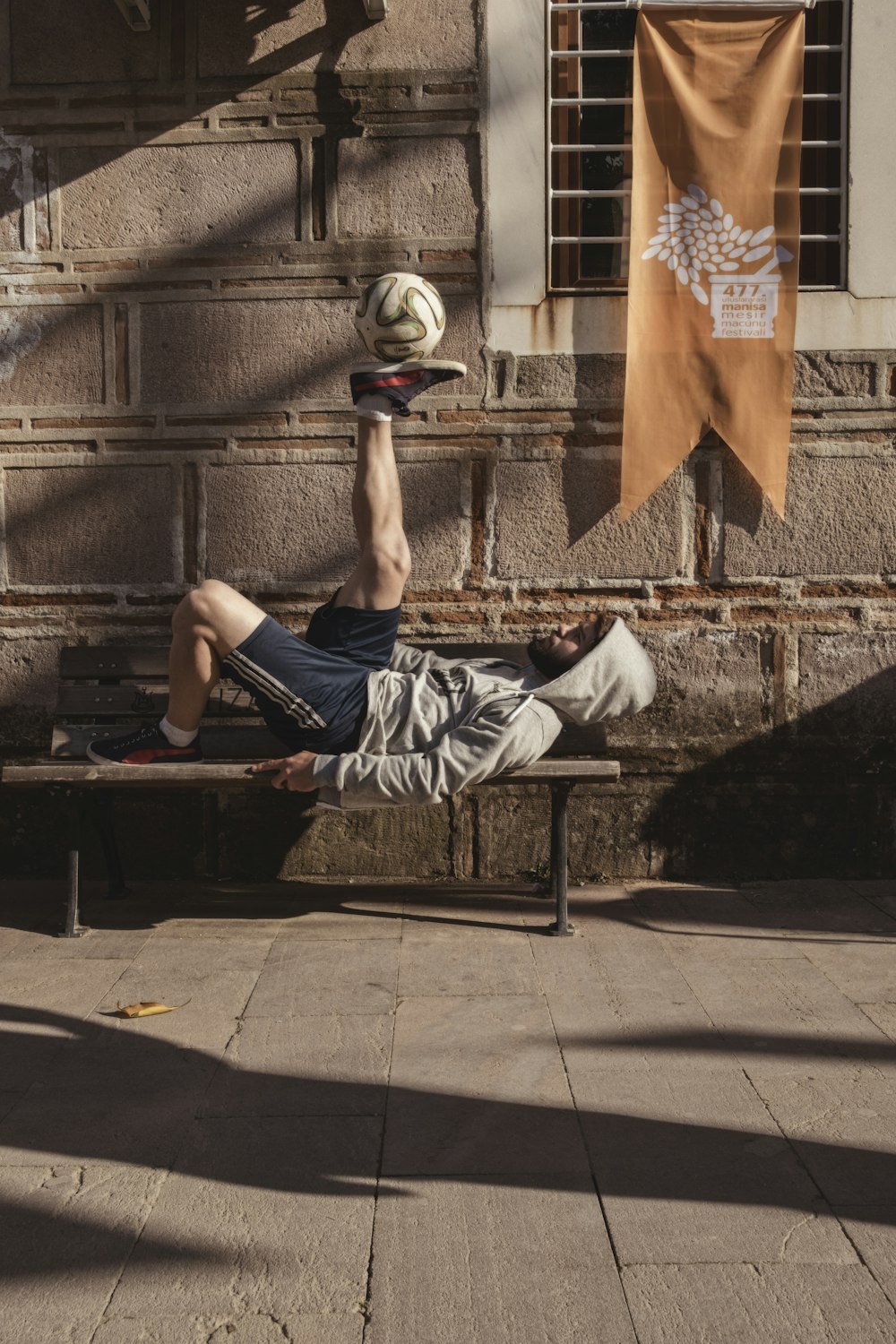 homem de jaqueta cinza com capuz e shorts pretos jogando bola enquanto deitado no banco
