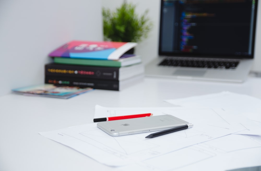 A smartphone on top of papers next to a laptop, books and pens.