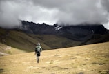A solitary hiker with a backpack and walking stick moves across a vast, barren landscape. The area is surrounded by majestic mountains partially obscured by heavy, dark clouds. The terrain is grassy and uneven, creating a dramatic and remote atmosphere.