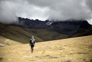 A solitary hiker with a backpack and walking stick moves across a vast, barren landscape. The area is surrounded by majestic mountains partially obscured by heavy, dark clouds. The terrain is grassy and uneven, creating a dramatic and remote atmosphere.