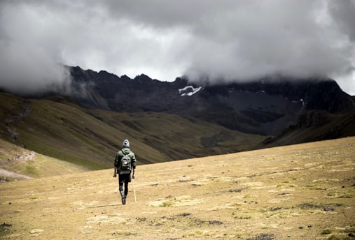A solitary hiker with a backpack and walking stick moves across a vast, barren landscape. The area is surrounded by majestic mountains partially obscured by heavy, dark clouds. The terrain is grassy and uneven, creating a dramatic and remote atmosphere.