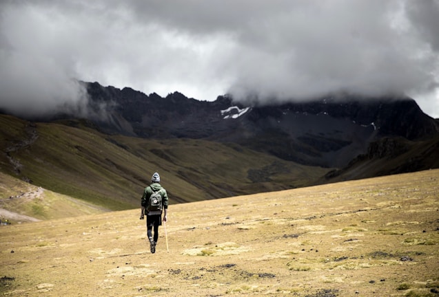 A solitary hiker with a backpack and walking stick moves across a vast, barren landscape. The area is surrounded by majestic mountains partially obscured by heavy, dark clouds. The terrain is grassy and uneven, creating a dramatic and remote atmosphere.