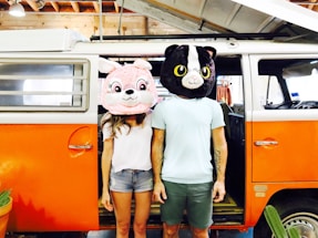 Two individuals are standing next to an orange and white van with its door open. They are wearing large, oversized animal mascot heads; one appears to be a pink rabbit and the other a black and white cat. The setting includes a rustic interior with wooden beams visible above.