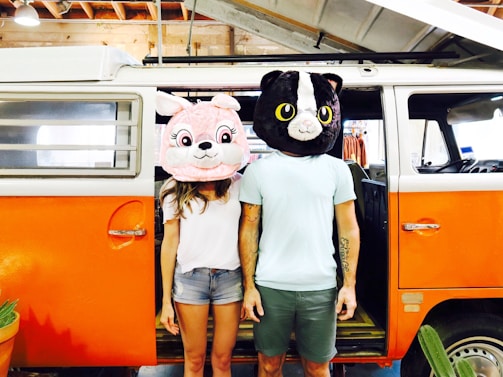 Two individuals are standing next to an orange and white van with its door open. They are wearing large, oversized animal mascot heads; one appears to be a pink rabbit and the other a black and white cat. The setting includes a rustic interior with wooden beams visible above.