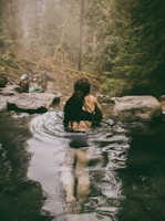 A person with long dark hair is relaxing in a natural hot spring surrounded by lush green forest. The setting is peaceful and serene, with steam rising from the water's surface. Large rocks are visible around the spring, and there is mist in the air, creating a mystical atmosphere.