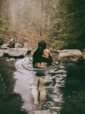A person with long dark hair is relaxing in a natural hot spring surrounded by lush green forest. The setting is peaceful and serene, with steam rising from the water's surface. Large rocks are visible around the spring, and there is mist in the air, creating a mystical atmosphere.
