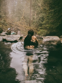 A person with long dark hair is relaxing in a natural hot spring surrounded by lush green forest. The setting is peaceful and serene, with steam rising from the water's surface. Large rocks are visible around the spring, and there is mist in the air, creating a mystical atmosphere.