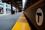 A train station platform with a train to the right, displaying a circular symbol with a T. The platform features yellow tactile paving and extends into the distance. Several pillars and the structure of the station roof are visible, giving a sense of depth.