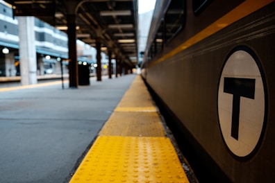 A train station platform with a train to the right, displaying a circular symbol with a T. The platform features yellow tactile paving and extends into the distance. Several pillars and the structure of the station roof are visible, giving a sense of depth.