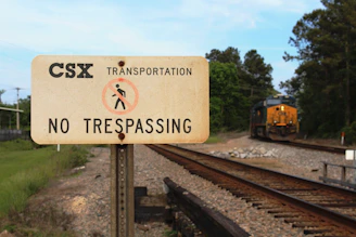 A CSX Transportation sign prominently features a 'No Trespassing' warning in black letters on a weathered beige background. In the distance, a CSX train travels along the railway tracks, surrounded by greenery and trees.