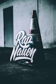A traffic cone sits on a dark surface in a dimly lit area, next to a sign with bold, stylized lettering that reads 'Rap Nation'. The background is industrial with a muted color palette.