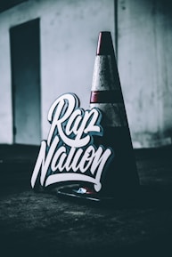 A traffic cone sits on a dark surface in a dimly lit area, next to a sign with bold, stylized lettering that reads 'Rap Nation'. The background is industrial with a muted color palette.