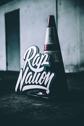 A traffic cone sits on a dark surface in a dimly lit area, next to a sign with bold, stylized lettering that reads 'Rap Nation'. The background is industrial with a muted color palette.