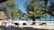A serene beach setting with a tranquil pool surrounded by lounge chairs. Palm trees and lush greenery provide shade, while the ocean stretches out in the background. Straw-roofed structures add to the tropical atmosphere.