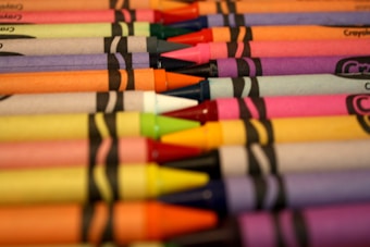 A collection of Crayola crayons arranged in parallel rows with their tips pointing to the right. The crayons feature vibrant colors including purple, orange, pink, blue, and green, and they are in their wrappers with the Crayola logo visible.