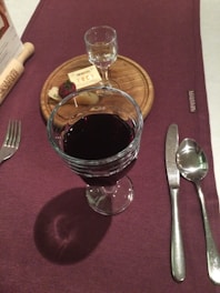 A dining table is set with a glass of red wine, a small clear glass of liquor on a wooden board, some cheese, a piece of bread, and a menu. The tablecloth is dark purple and there is cutlery neatly arranged next to a napkin.
