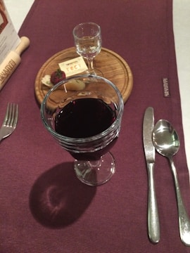 A dining table is set with a glass of red wine, a small clear glass of liquor on a wooden board, some cheese, a piece of bread, and a menu. The tablecloth is dark purple and there is cutlery neatly arranged next to a napkin.