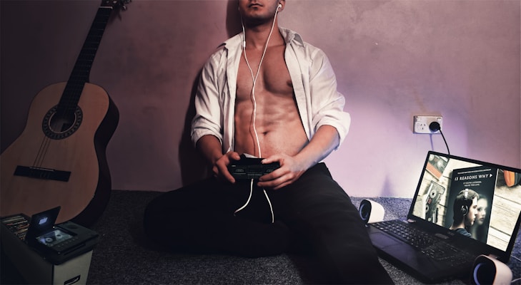 A shirtless person is sitting on the floor, wearing earphones, and holding a game controller. A classical guitar is leaning against the wall nearby, and on the other side is an open laptop displaying a show on its screen. A box filled with CDs is next to the guitar.
