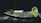 A slice of green tea cake sits on a dark plate, dusted with powdered sugar and surrounded by a sprinkle of matcha powder. In the background, a sieve is seen tipped over with some of the matcha powder spilled onto the surface.