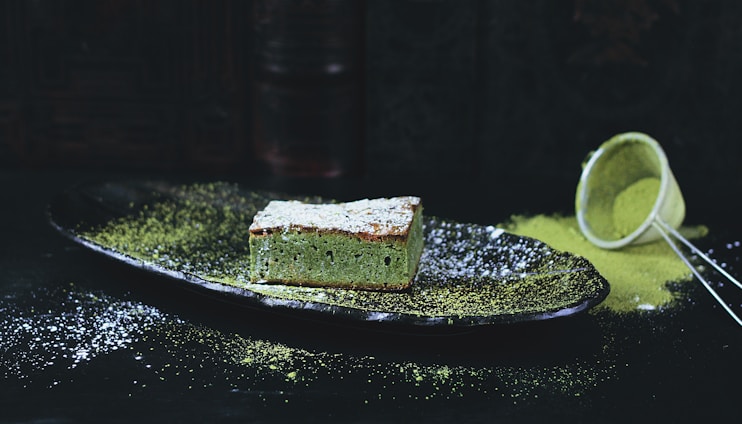 A slice of green tea cake sits on a dark plate, dusted with powdered sugar and surrounded by a sprinkle of matcha powder. In the background, a sieve is seen tipped over with some of the matcha powder spilled onto the surface.