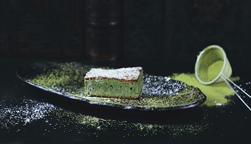 A slice of green tea cake sits on a dark plate, dusted with powdered sugar and surrounded by a sprinkle of matcha powder. In the background, a sieve is seen tipped over with some of the matcha powder spilled onto the surface.