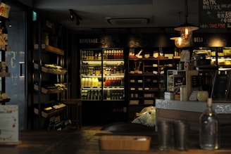 A cozy interior of a small market or cafe featuring wooden shelves stocked with wine bottles and a refrigerated section filled with various beverages. Dim lighting creates a warm and inviting atmosphere. There is a deli counter with food items and overhead hanging lights that add to the ambiance.