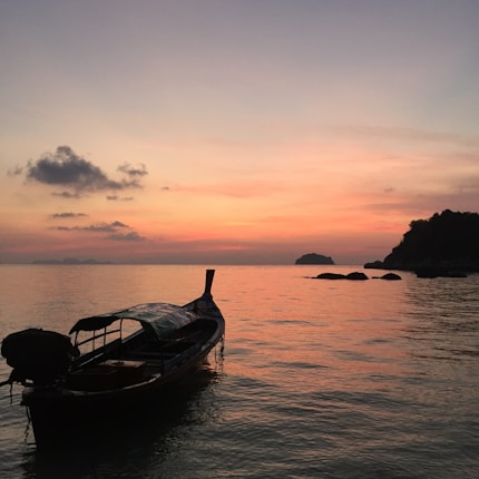 A serene sunset over a calm sea with a long-tail boat at the forefront and silhouetted trees and rocks in the distance. The sky is painted with gradient hues of orange, pink, and purple, creating a tranquil and picturesque scene.